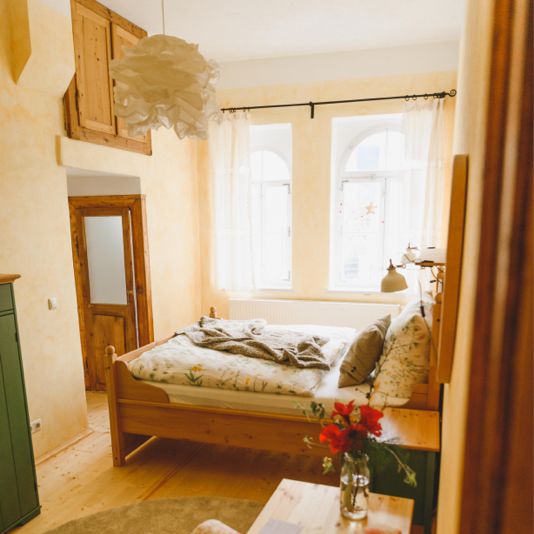 Helles Schlafzimmer mit Doppelbett, gelb gestrichen. Im Vordergrund ein Sessel mit Blumenmuster.