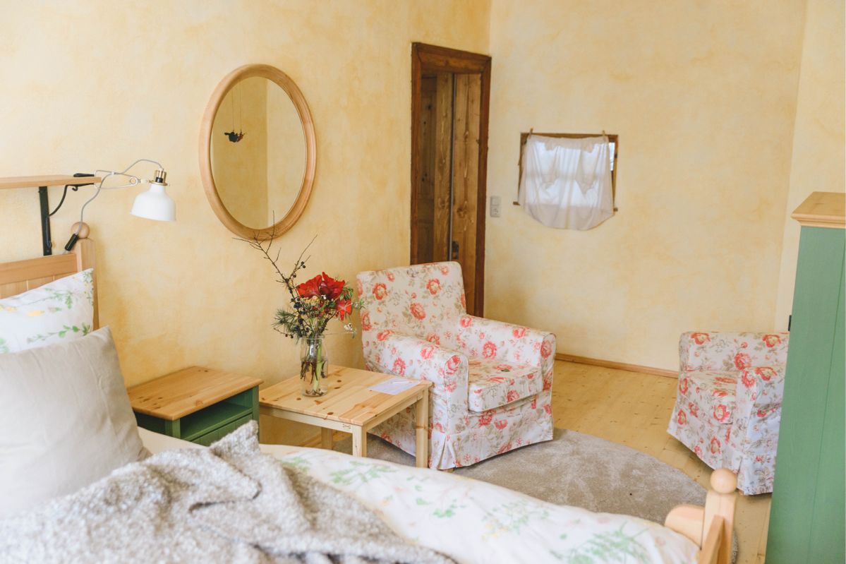 gelb gestrichenes Schlafzimmer, im Vordergrund ein Bett, im Hintergrund zwei Sessel mit Blumenmuster sowie Tisch mit frischen Blumen