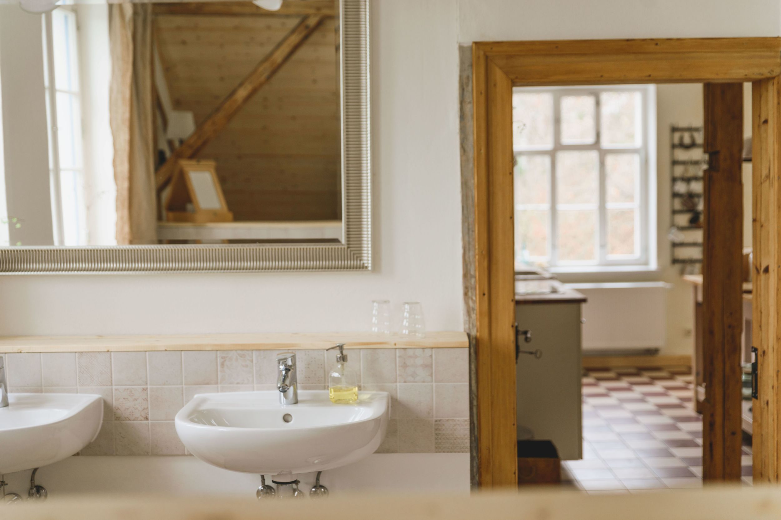 Blick vom Badezimmer in die Wohnküche. Im Vordergrund zwei Waschbecken und ein großer Spiegel, im Hintergrund eine offene Tür zur Küche.
