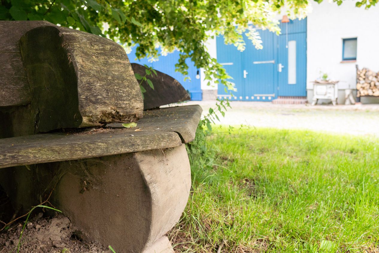 Detailaufnahme rustikale Sitzbank um Stamm der Linde im Innenhof, im Hintergrund weiteres Gebäude und Holzstapel
