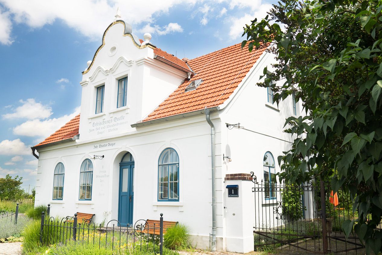 Ferienhaus Alte Wasserschänke Totale Ferienhaus: weißes Haus mit rotem Ziegeldach und blauer Tür, davor zwei Bänke im Grünen.