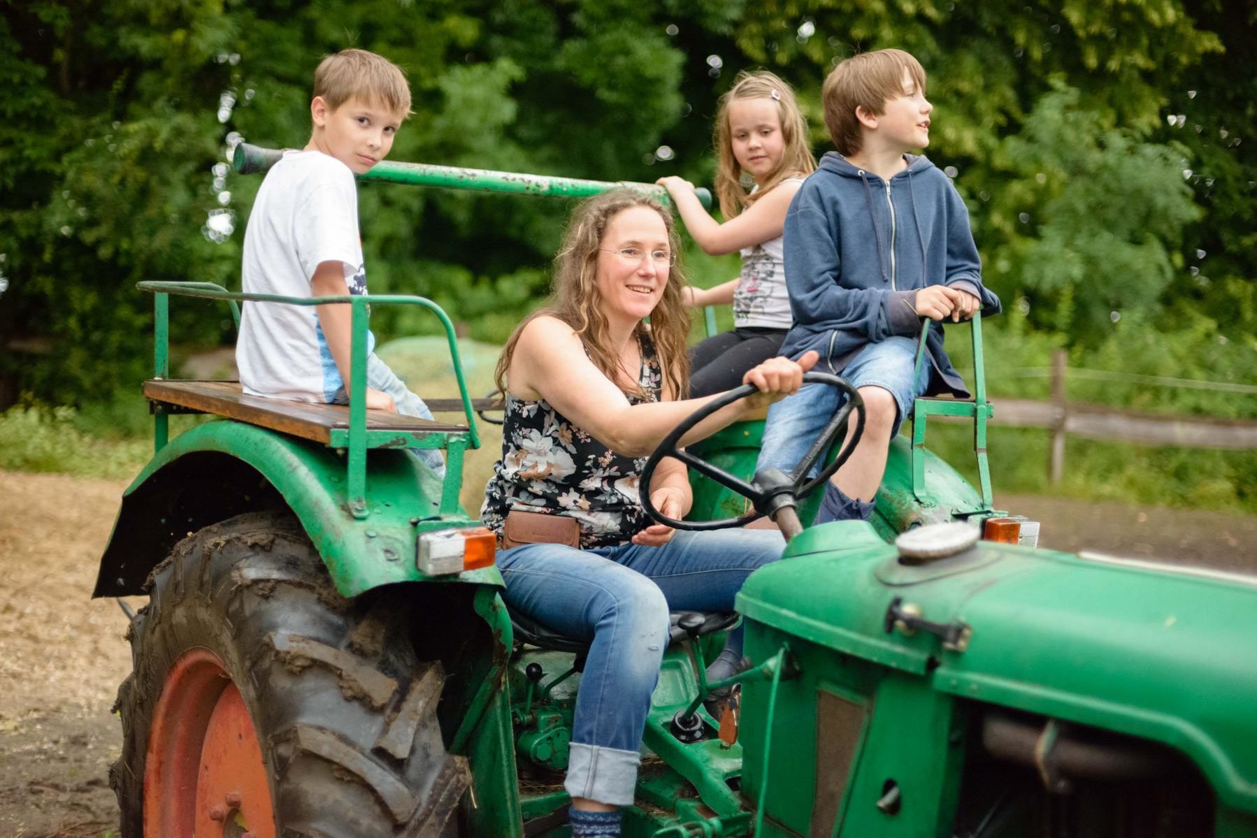 Frau fährt grünen Traktor, hinter ihr sitzen drei Kinder mit auf dem Traktor, zwei Jungen und ein Mädchen