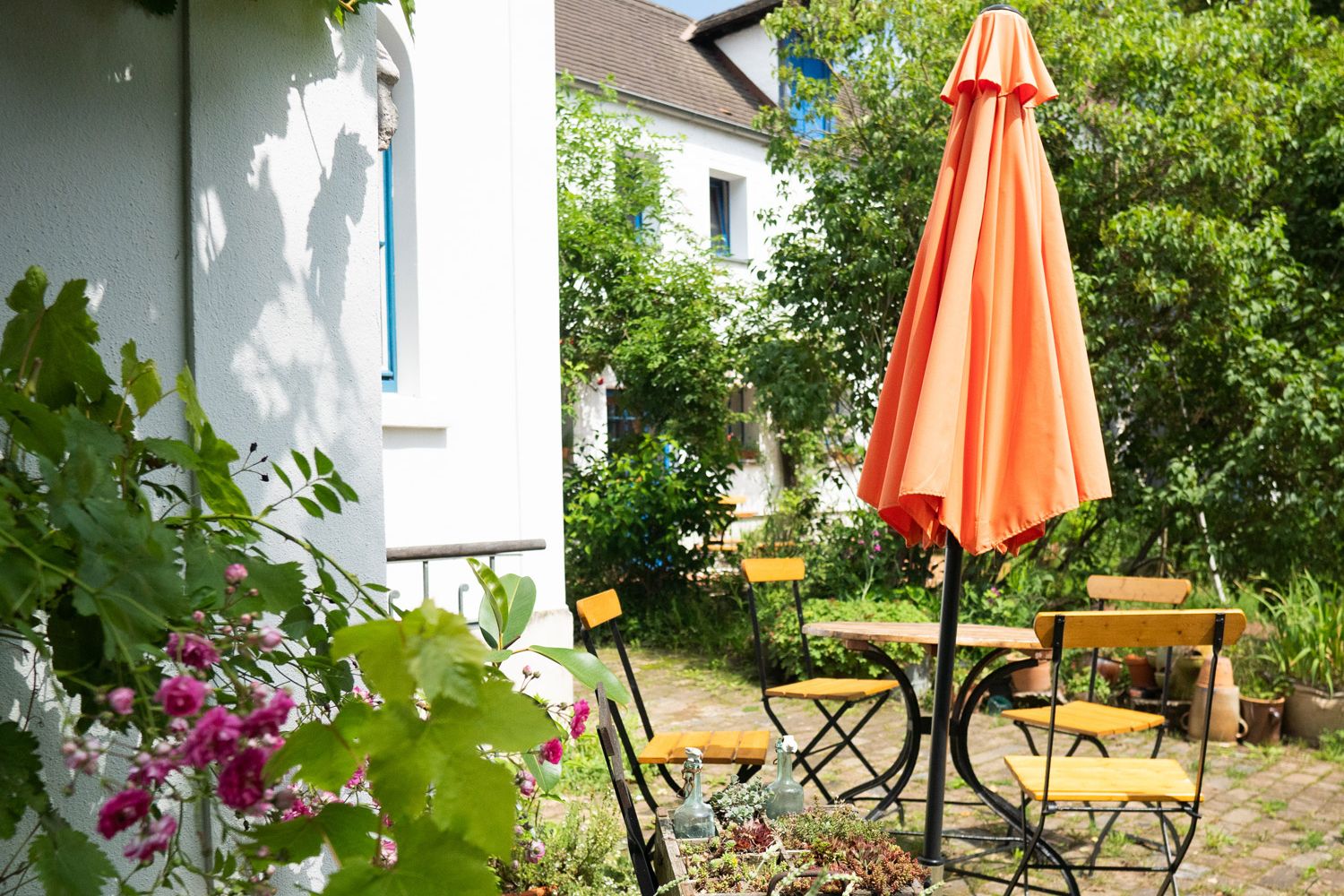 Sitzbereich im Grünen mit Tisch, vier Stühlen und Sonnenschirm. Im Hintergrund Haus sowie grüne Bäume und Sträucher.
