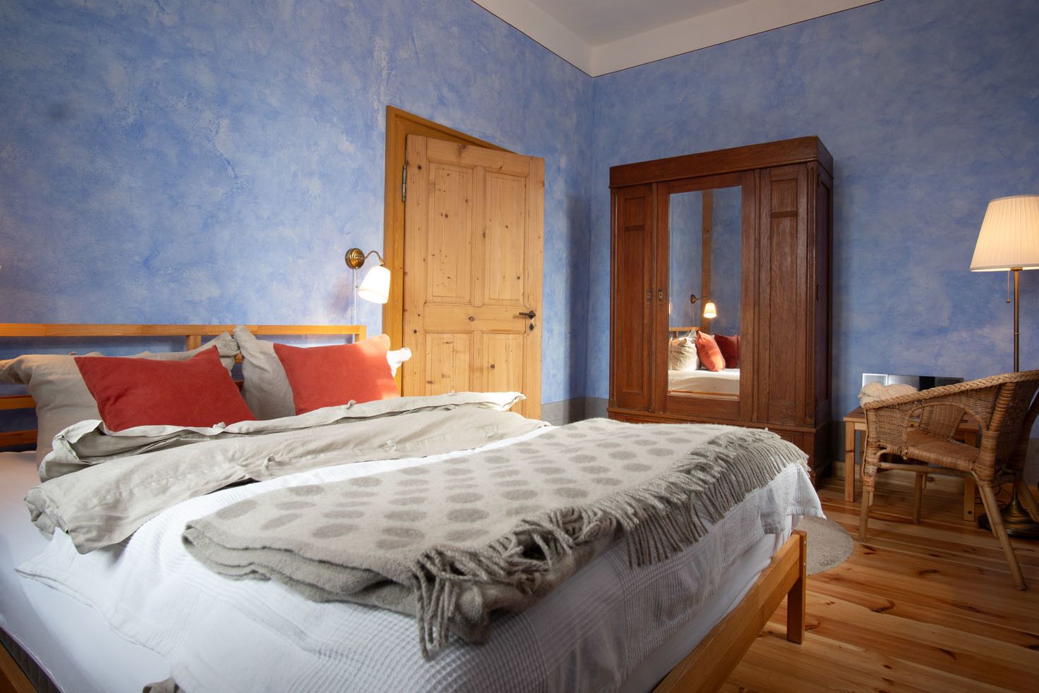 blau gestrichenes Schlafzimmer, Blick aus der Zimmerecke auf Tür und Kleiderschrank mit Spiegel, in dem sich das Bett mit vielen Kissen spiegelt